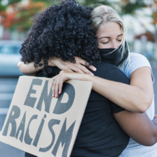 Een vrouw met lichte huidskleur en een bord 'End racism' in haar hand omarmt een vrouw met donkere huidskleur.